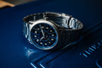 SLOMO Watches Glance automatic watch featuring a blue dial, white Arabic numerals, and brushed steel bracelet resting with the clasp visible.