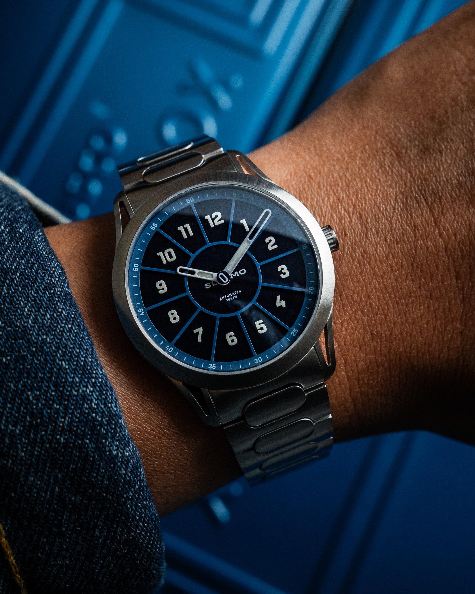SLOMO Watches Glance automatic watch on wrist featuring a blue dial, white Arabic numerals, orange hands, and a brushed steel bracelet.