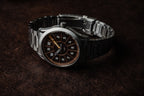 SLOMO Watches Glance automatic watch featuring a brown dial, white Arabic numerals, and brushed steel bracelet resting with the clasp visible.