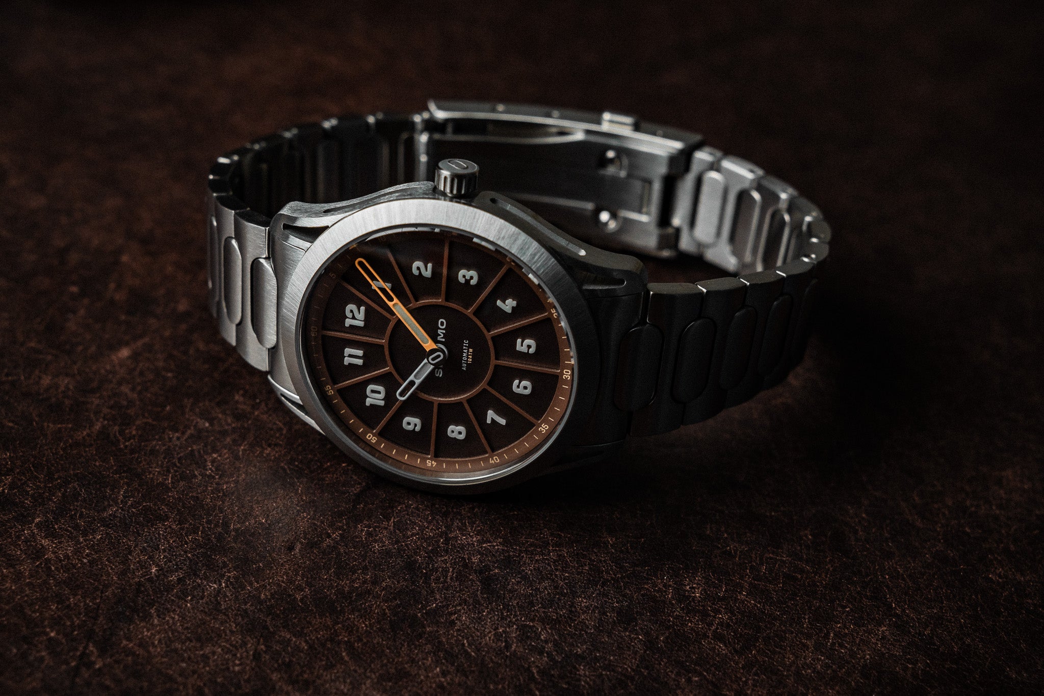 SLOMO Watches Glance automatic watch featuring a brown dial, white Arabic numerals, and brushed steel bracelet resting with the clasp visible.
