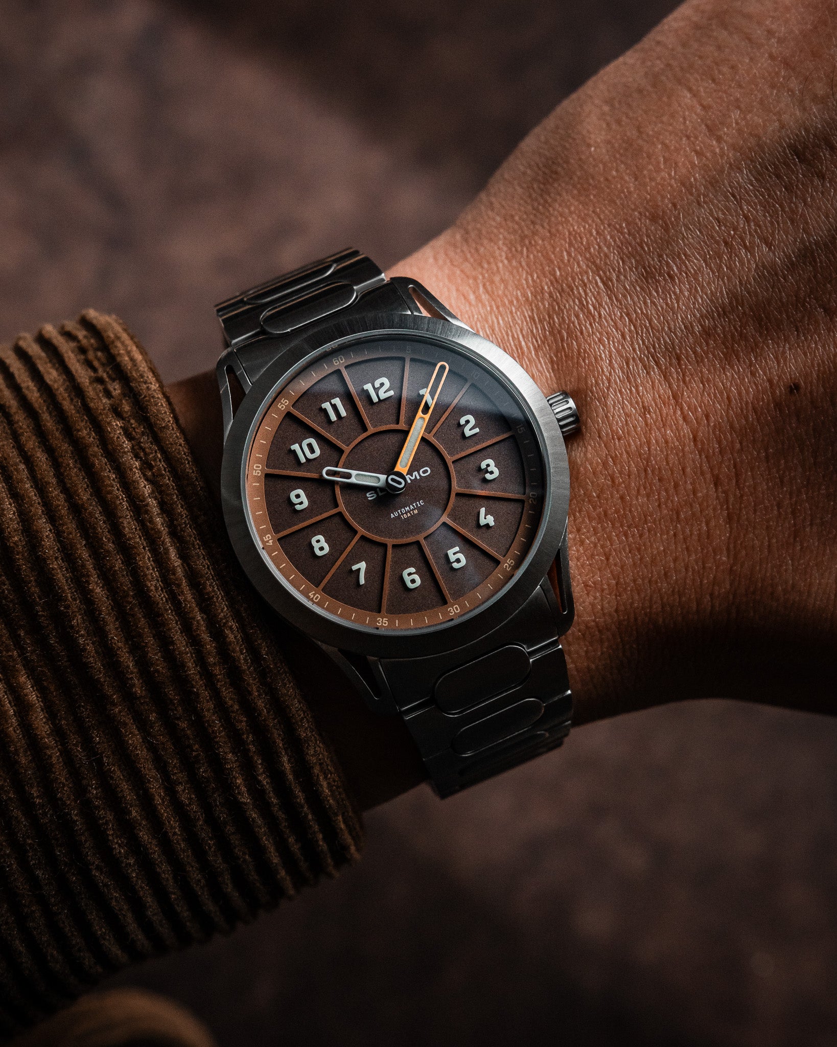 SLOMO Watches Glance automatic watch on wrist featuring a brown dial, white Arabic numerals, orange hands, and a brushed steel bracelet.