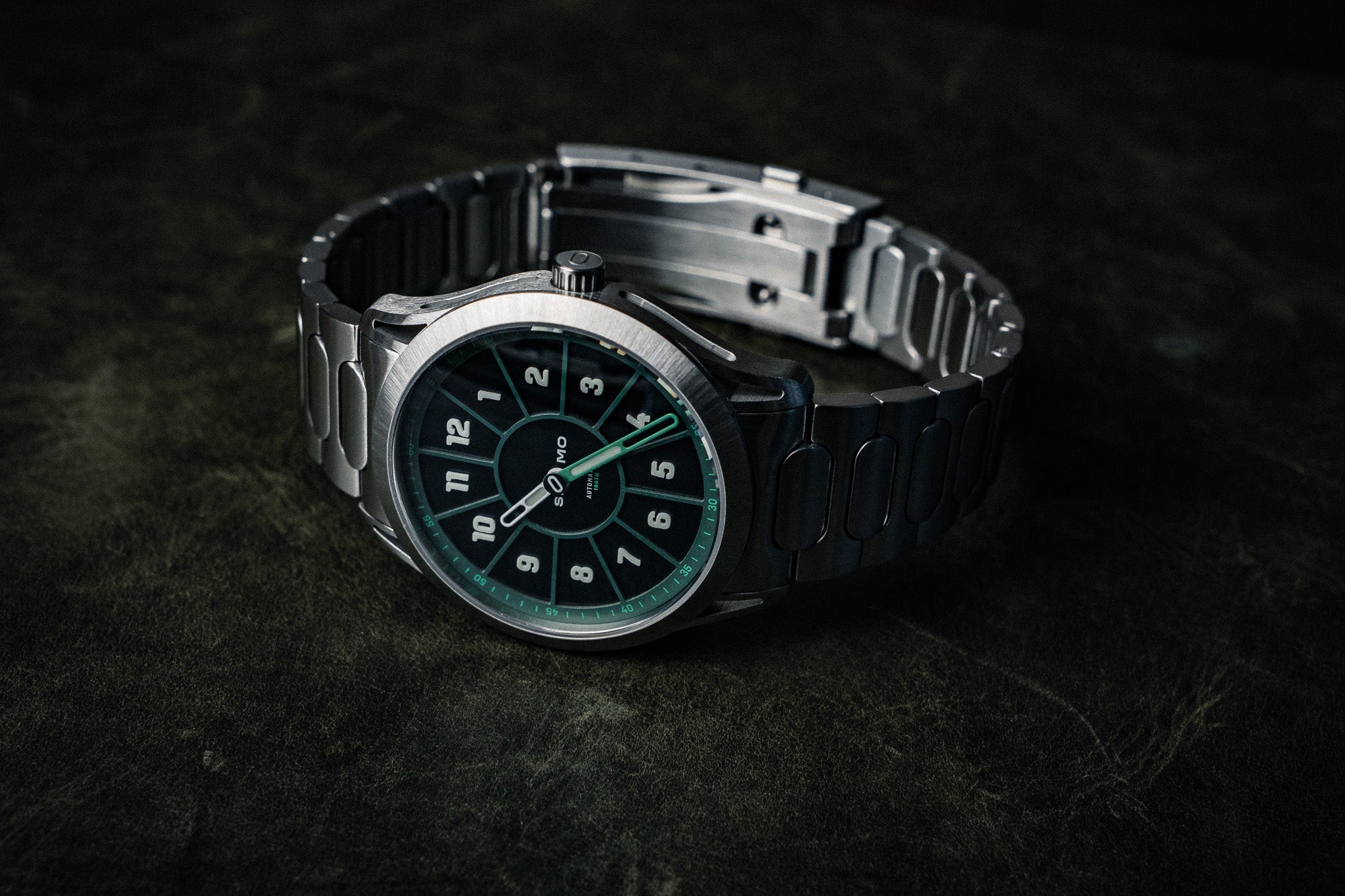 SLOMO Watches Glance automatic watch featuring a green dial, white Arabic numerals, and brushed steel bracelet resting with the clasp visible.