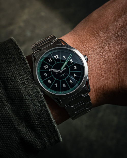 SLOMO Watches Glance automatic watch on wrist featuring a green dial, white Arabic numerals, orange hands, and a brushed steel bracelet.
